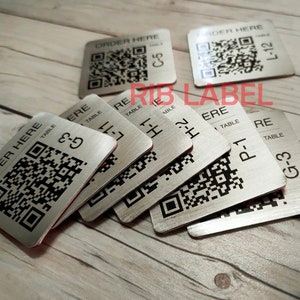 Puede incluir: Primer plano de varias etiquetas de número de mesa de metal plateado con un código QR negro y el texto "ORDER HERE" y el número de mesa. Las etiquetas están sobre una superficie de madera.