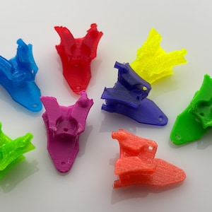 May include: A collection of seven 3D-printed objects in various bright colors, including blue, red, yellow, purple, green, and orange. The objects have a unique, abstract shape, and are arranged on a white surface.