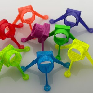 May include: A collection of colorful, 3D-printed plastic objects in various hues, including red, orange, purple, green, blue, and yellow. Each object has a ring-shaped center with three legs and a small, rounded foot.