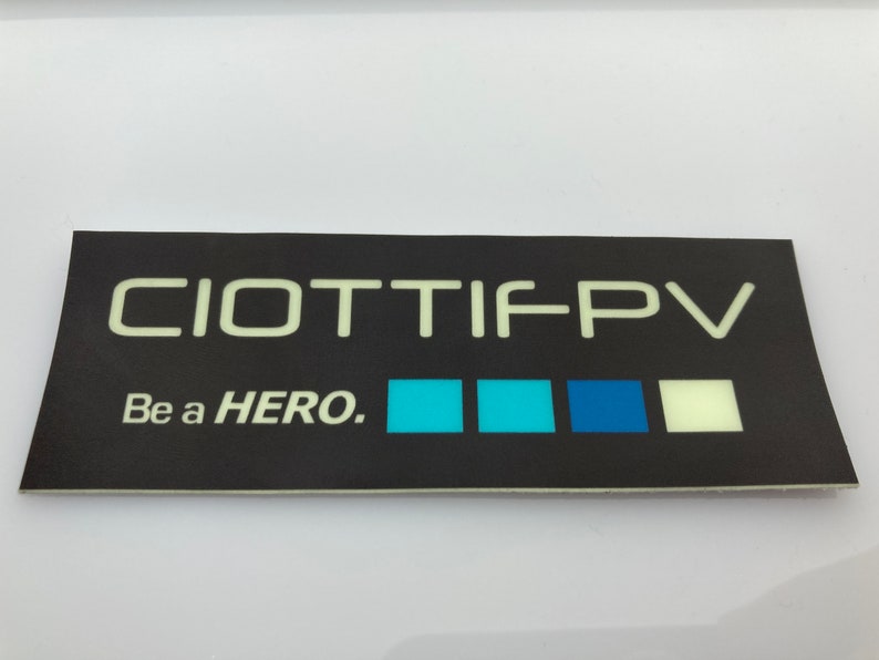 May include: Black and white sticker with the text "CIOTTIFPV" and "Be a HERO."  The sticker has a row of four colored squares: light blue, turquoise, dark blue, and white.