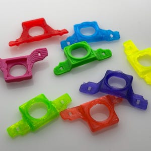 May include: A collection of colorful, 3D-printed plastic components in various hues, including red, pink, blue, green, yellow, and purple. Each piece features a circular opening and a unique, geometric design, suggesting a functional or decorative purpose.