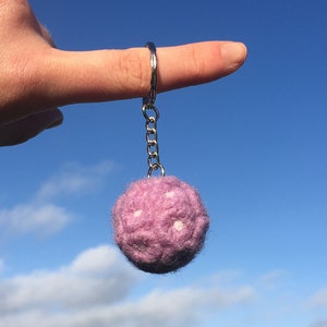 Op de afbeelding: Een sleutelhanger met een lavendelkleurige vilten bal met witte accenten, afgebeeld tegen een helderblauwe lucht. De sleutelhanger is bevestigd aan een zilveren ring en ketting. De achtergrond toont een groen veld en bomen.