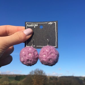 May include: Two lilac felt ball earrings with white spots, suspended from a black tag. The tag has the word "nudijai" in white. The backdrop is a clear blue sky and a green landscape, typical of the British countryside.