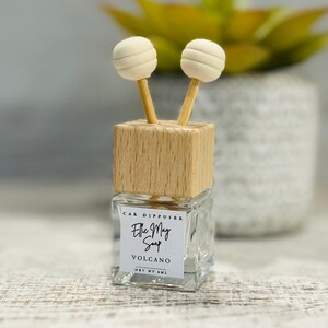 Könnte beinhalten: Eine kleine Glasflasche mit einem Holzdeckel und zwei weißen Diffusor-Kugeln. Die Flasche hat ein Etikett mit der Aufschrift "Car Diffuser Ellie May Soap Volcano Net Wt 8mL".