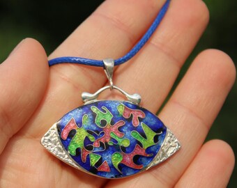 Puede incluir: Un collar con colgante azul con un engaste plateado y un cordón azul. El colgante tiene un fondo azul con formas abstractas coloridas en verde, rosa y amarillo. El collar se sostiene en una mano.