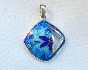 Puede incluir: Un colgante de plata con un diseño de esmalte azul y morado que presenta una flor estilizada. El colgante tiene forma de diamante con un recorte en un lado.