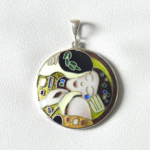 Puede incluir: Un colgante redondo de plata con un diseño de esmalte colorido que representa a una pareja abrazándose. El diseño incluye una mujer con un sombrero negro y un hombre con una corona de hojas verdes. Los colores del esmalte son negro, blanco, verde, amarillo, azul y rojo.