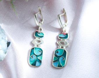 May include: A pair of silver dangle earrings with teal and blue enamel designs. The earrings have a rectangular bottom shape and a curved top, with a series of oval and rectangular shapes. Pink flowers and green leaves are in the background.