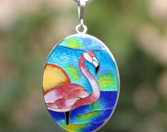 Op de afbeelding: Een zilveren ketting met een ovale hanger met daarop een roze flamingo die in blauw water staat met een gele zon op de achtergrond. De hanger is versierd met kleurrijk email.