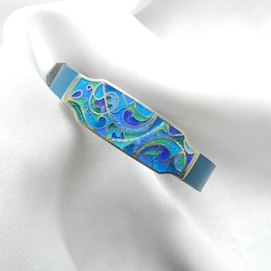 Puede incluir: Un brazalete de cuero azul con una placa rectangular de plata con un dise&ntilde;o de esmalte colorido de espirales azules, verdes y dorados.