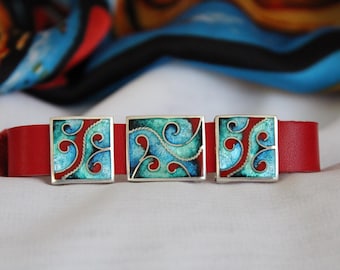 Puede incluir: Tres gemelos de plata con diseños de esmalte azul y rojo. Los gemelos están colocados sobre una correa de cuero roja.
