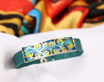 Puede incluir: Una pulsera de cuero turquesa con un dije rectangular plateado con un diseño de espiral azul, verde y amarillo.