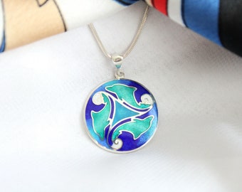 Puede incluir: Un collar de plata con un colgante redondo de esmalte azul que presenta tres delfines estilizados en tonos de azul y verde.