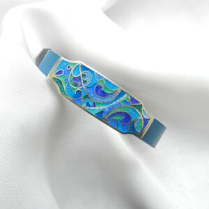 Puede incluir: Un brazalete de cuero azul con una placa rectangular de plata con un dise&ntilde;o de esmalte colorido de espirales azules, verdes y dorados.