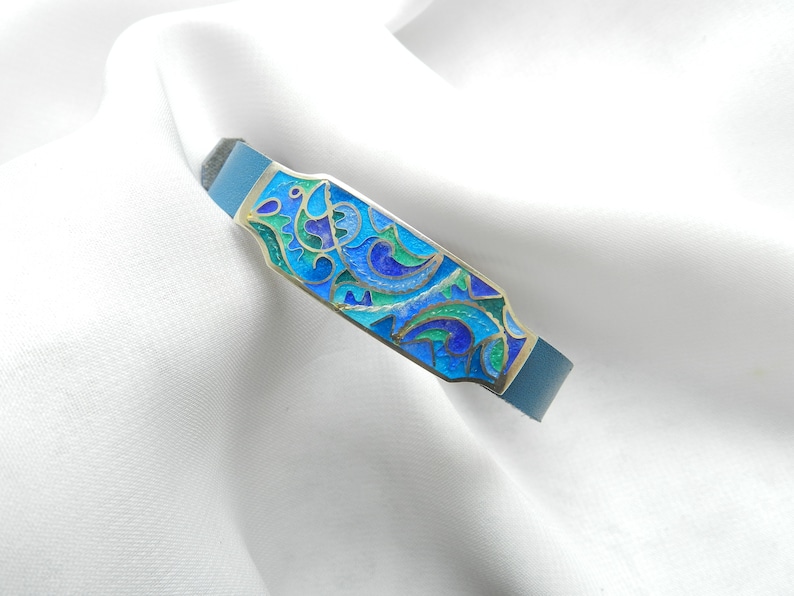 Puede incluir: Un brazalete de cuero azul con una placa rectangular de plata con un dise&ntilde;o de esmalte colorido de espirales azules, verdes y dorados.