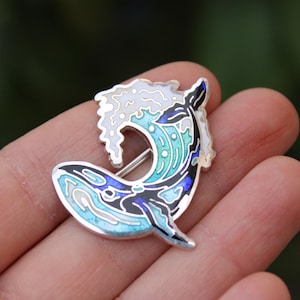 May include: A silver-toned whale pin with enamel detailing. The pin features a whale leaping through a wave, with shades of blue, black, and white. The pin is a decorative accessory.