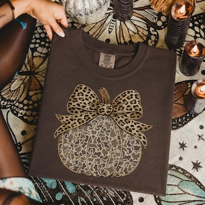 Op de afbeelding: Een donkerbruin t-shirt met een afbeelding van een pompoen met een lichtbruin en grijs koeienprintpatroon. De pompoen is bekroond met een grote strik met een beige en zwart luipaardprintpatroon. Het shirt heeft een Comfort Colors label.