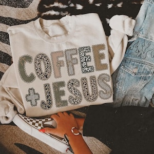 Puede incluir: Una sudadera de cuello redondo color crema con el texto COFFEE + JESUS en letras grandes con bordes festoneados. Las letras están rellenas con varios patrones, incluyendo estampado de leopardo, rayas blancas y negras y verde salvia.