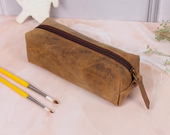 May include: A brown leather pencil case with a zipper closure and a leather pull tab. Two yellow paintbrushes with white bristles are in the foreground. The pencil case is rectangular and has a distressed finish.