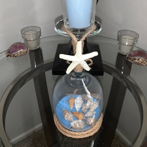 May include: A glass candle holder with a blue votive candle, a starfish, and seashells inside. The holder is wrapped with twine and sits on a glass table.