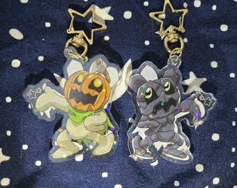 Puede incluir: Dos llaveros de acrílico transparente con cierres dorados en forma de estrella. Uno presenta un personaje de dibujos animados con cabeza de calabaza y el otro un personaje gris con aspecto de robot. Ambos tienen detalles brillantes y están sobre fondo azul oscuro.