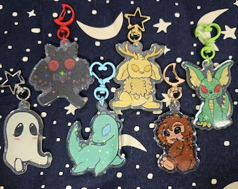 Puede incluir: Una colección de llaveros acrílicos con varias criaturas de dibujos animados. Los llaveros incluyen un fantasma, un dinosaurio, una polilla, una criatura parecida a un conejo, un bigfoot y un monstruo parecido a un dragón. Cada llavero tiene un cierre de diferente color.