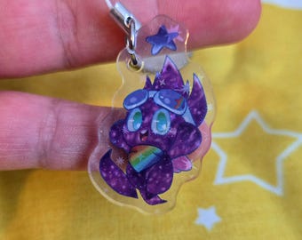 Puede incluir: Un dije acrílico transparente con un personaje de dibujos animados púrpura con un diseño de estrellas, que lleva gafas y un sombrero de mago. El personaje sostiene un corazón arcoíris. Un pequeño dije de estrella cuelga arriba.