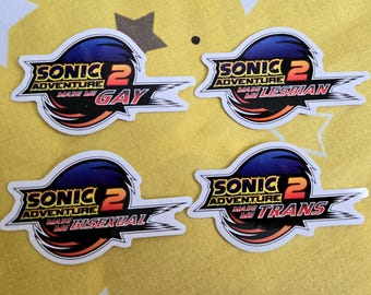 Puede incluir: Cuatro pegatinas con temática de Sonic the Hedgehog. Cada pegatina tiene un gráfico azul y negro con el texto "SONIC 2 ADVENTURE MADE ME GAY", "SONIC 2 ADVENTURE MADE ME LESBIAN", "SONIC 2 ADVENTURE MADE ME BISEXUAL" y "SONIC 2 ADVENTURE MADE ME TRANS".