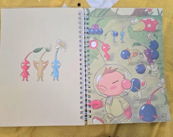 Puede incluir: Cuaderno de espiral con ilustraciones coloridas de personajes de la serie de videojuegos Pikmin. La obra de arte incluye un personaje con traje espacial y varios Pikmin que llevan frutas azules y moradas. El cuaderno está abierto, mostrando la obra.