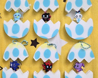 Puede incluir: Nueve llaveros con personajes de dibujos animados dentro de diseños de cáscaras de huevo. Las cáscaras de huevo son blancas con lunares azules y una parte superior amarilla. Cada llavero tiene una cadena plateada y un personaje diferente en el interior.
