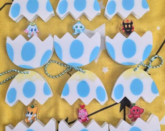 Puede incluir: Seis llaveros de acrílico con forma de cáscara de huevo blanca con puntos azules, cada uno con una pequeña figura en su interior. Los llaveros están unidos a cadenas de bolas plateadas y muestran varios personajes de dibujos animados.