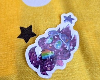 Puede incluir: Una pegatina morada con un personaje de dibujos animados con elementos espaciales. El personaje tiene ojos grandes, un acento arcoíris y está rodeado de estrellas. La pegatina está sobre un fondo amarillo con una luna creciente blanca y una estrella negra.