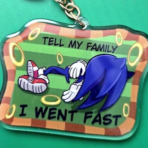 May include: A blue and white acrylic keychain featuring a cartoon character lying down with the text "Tell my family I went fast".