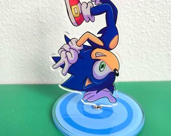 Puede incluir: Un personaje de dibujos animados azul y amarillo, Sonic the Hedgehog, está de pie sobre una base de remolino azul y blanco. El personaje está boca abajo con sus zapatos rojos en el aire.