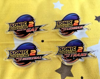 Puede incluir: Cuatro pines acrílicos transparentes con el logo de Sonic 2 Adventure, cada uno con una palabra diferente: "GAY", "LESBIAN", "BISEXUAL" y "TRANS". Los pines presentan un diseño azul y naranja con detalles en blanco y negro.