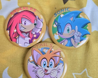 Puede incluir: Tres botones redondos con personajes de la franquicia Sonic the Hedgehog. Los botones muestran a Knuckles, Sonic y Tails, cada uno con sus nombres sobre sus imágenes. Los botones están sobre un fondo amarillo.