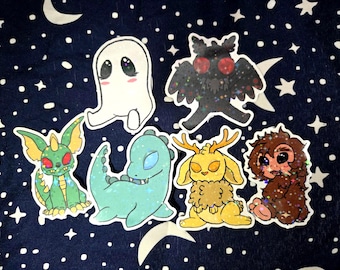 Puede incluir: Una colección de pegatinas de criaturas caprichosas, que incluyen un fantasma blanco, una polilla negra, un dragón verde, un dinosaurio verde azulado, una criatura amarilla parecida a un conejo y una criatura marrón y peluda. Cada pegatina tiene un borde blanco.