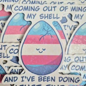 May include: A die-cut sticker featuring an egg with a cracked top, colored in horizontal stripes of light blue, pink, and white. The egg has a simple smiling face and the text "COMING OUT OF MY SHELL AND I'VE BEEN DOING JUST FINE" in blue.
