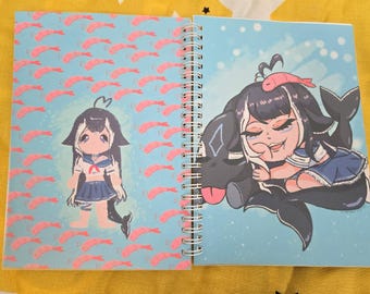 Puede incluir: Cuaderno de espiral con una cubierta azul con ilustraciones de estilo anime. Una página muestra un personaje con uniforme de marinero con un fondo de patrón de camarones. La otra página representa un personaje con un pez en la cabeza, descansando sobre una criatura negra.