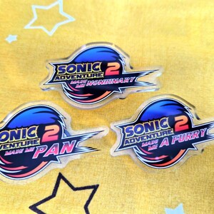May include: Three acrylic pins featuring a stylized logo design inspired by Sonic Adventure 2. The text on the pins reads Sonic Adventure 2 Made Me Nonbinary, Sonic Adventure 2 Made Me Pan, and Sonic Adventure 2 Made Me A Furry in blue, red, and pink.