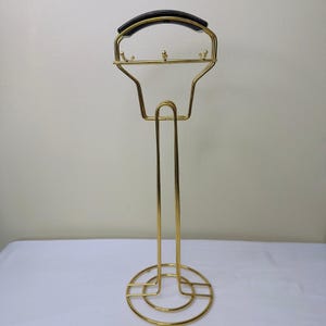 Könnte beinhalten: Ein goldfarbener Metall-Herrendiener mit schwarzem Griff. Der Ständer hat einen kreisförmigen Fuß und eine geschwungene Oberseite mit kleinen Haken zum Aufhängen von Gegenständen. Das Design ist schlicht und minimalistisch.
