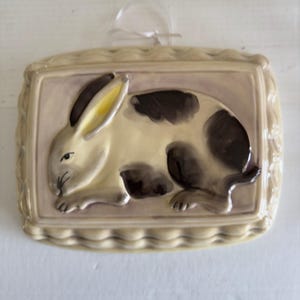 May include: A ceramic wall hanging featuring a rabbit design. The rabbit is cream-colored with dark brown spots and black ears. The background is a light purple, and the frame is a light beige with a decorative edge. A clear ribbon is attached for hanging.