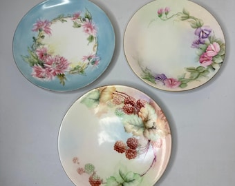 May include: Three round ceramic plates featuring hand-painted botanical designs. One plate has a light blue background with pink flowers, another has a cream background with purple and pink flowers, and the third features red berry clusters.