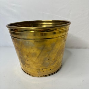 Pode incluir: Um vaso de latão polido com formato cilíndrico cônico e uma parte superior larga e aberta. A superfície metálica apresenta um acabamento dourado quente com variações sutis de tom e pequenas manchas escuras no corpo.