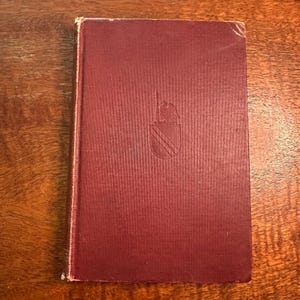 May include: A hardcover book with a deep maroon fabric cover. The front cover features a centered, embossed heraldic emblem depicting a shield with diagonal lines and a small bird figure perched on top. The book has a vertical spine on the left side.