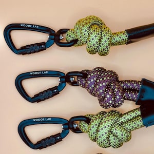 Puede incluir: Tres correas para perros con mosquetones negros y cuerda anudada. Los mosquetones tienen el texto "WOOF LAB" impreso. La cuerda es verde y morada, y el fondo es de color naranja claro.