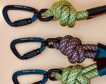 May include: Three dog leads with black carabiner clips and knotted rope. The carabiners have the text "WOOF LAB" printed on them. The rope is green and purple, and the background is a light orange colour.