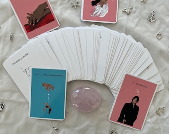 May include: A spread of tarot cards with pink, blue, and white backgrounds. The cards feature illustrations of people and symbols. The cards are laid out in a fan shape on a white surface. There are several small silver charms scattered around the cards.