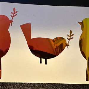 May include: Three stylized bird silhouettes with olive branches in their beaks. The birds are red, green, and yellow with a metallic iridescent finish.