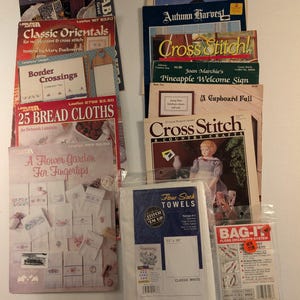 May include: A collection of cross-stitch pattern booklets and instructional leaflets, including titles like 25 Bread Cloths, A Flower Garden for Fingertips, and Autumn Harvest, alongside a package of flour sack towels and a Bag-It floss organizer system.